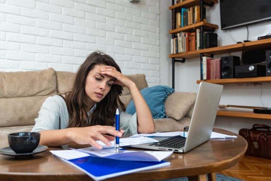 Stressed young woman sitting at a coffee table at home, reviewing bills with an open laptop and a mug nearby