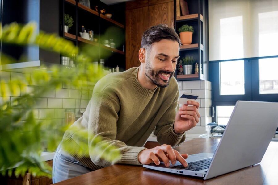 Smiling man using his laptop in the kitchen to pay his credit card
