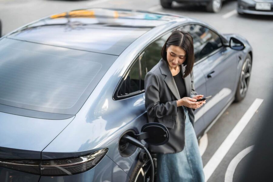 Young woman standing beside a parked electric car, holding a smartphone while the vehicle is connected to a charging cable, with other cars and a city street visible in the background.