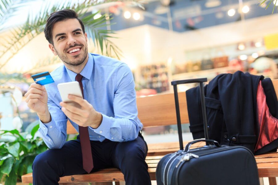 Young man using smart phone and credit card, next to him a suitcase.