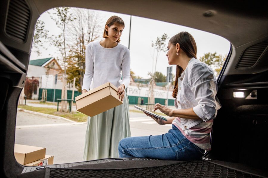Candid shot of young woman sitting in a car trunk and using digital tablet, checking her auto insurance and adding her roommate as an excluded driver.