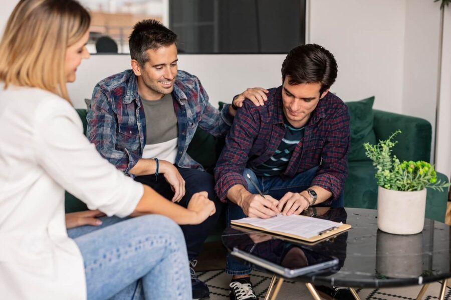 Young man signing a lease, with a guarantor beside him. A female real estate agent sits nearby.