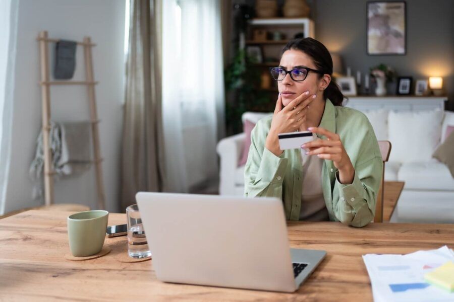 Concerned young woman sitting at her desk at home and holding a credit card, with an open laptop and a glass of water beside her