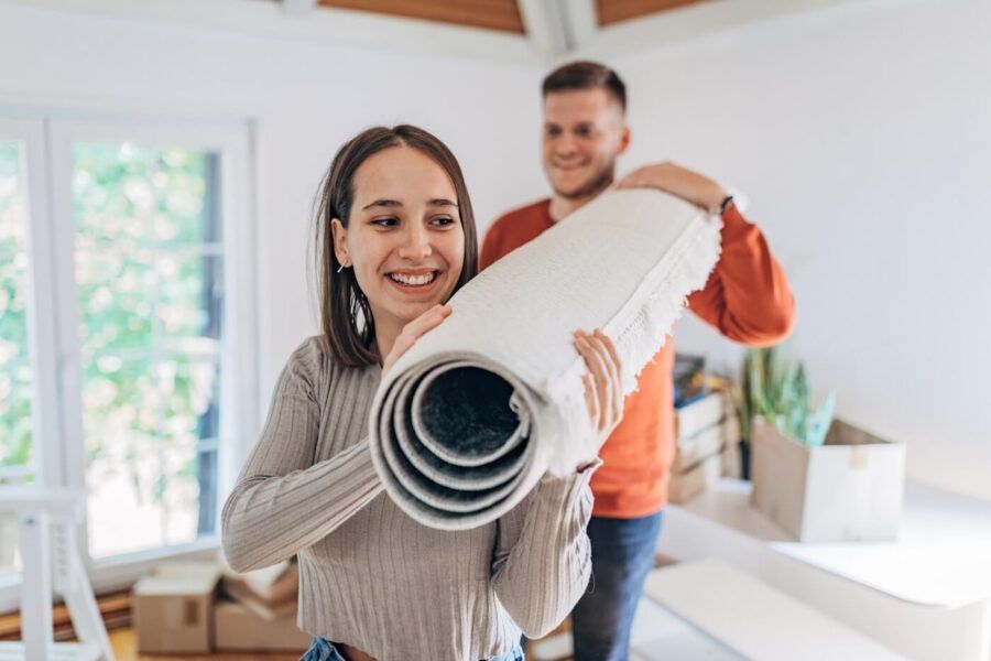Smiling young couple carrying a rolled-up carpet into their new apartment, surrounded by moving boxes.