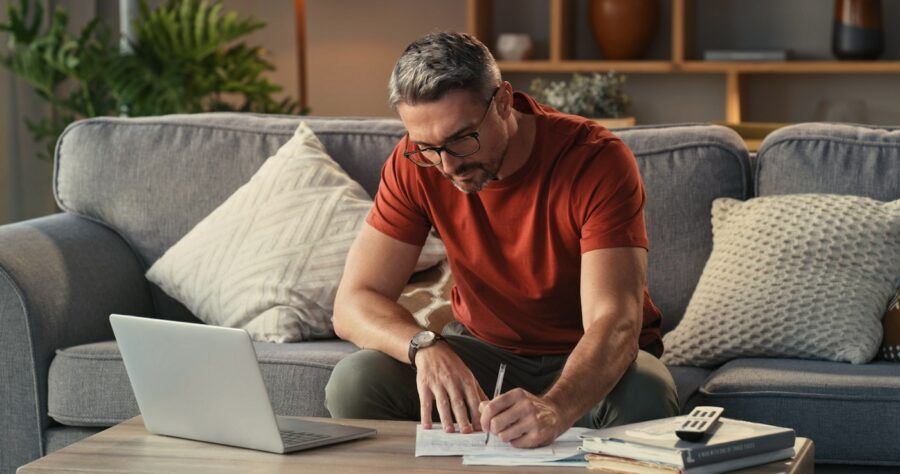 Shot of a mature man using a laptop and going through paperwork on the sofa at home