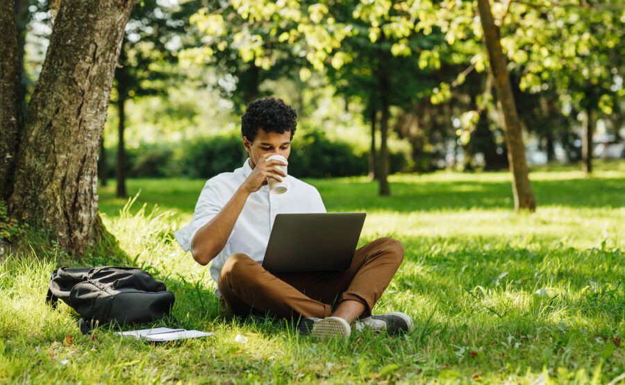 Man drinking coffee, sitting in the grass while working on laptop in the park