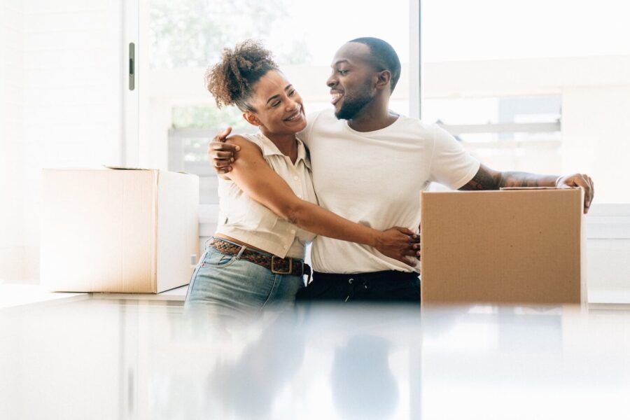 Happy couple hugging each other in a new home; the man is holding a moving box with one hand.