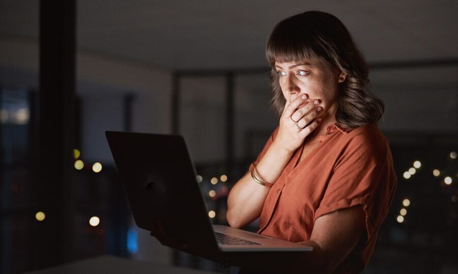 A shocked woman covers her mouth with one hand while staring at a laptop screen in a dimly lit office