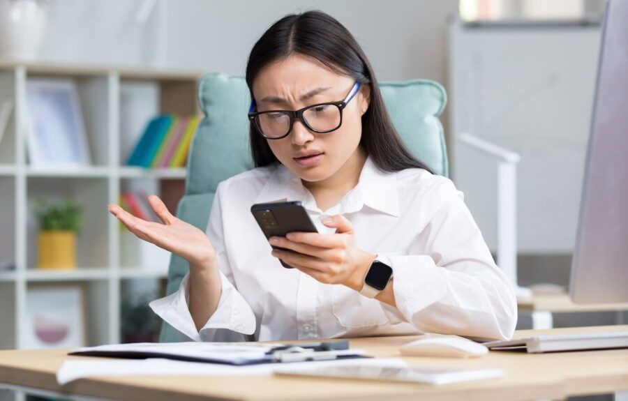 Concerned woman wearing glasses, looking at her smartphone while seated at a desk in an office setting