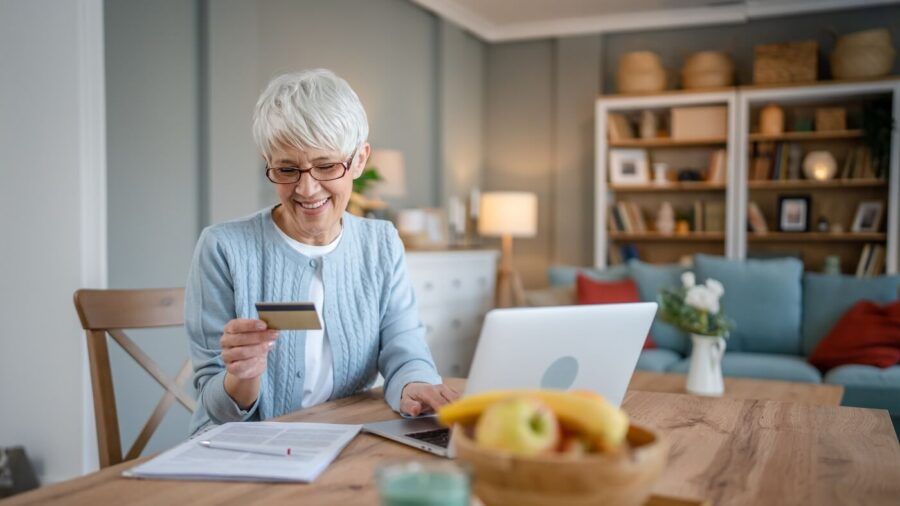 Senior woman sitting at a table, using her laptop to make an online payment with a debit card