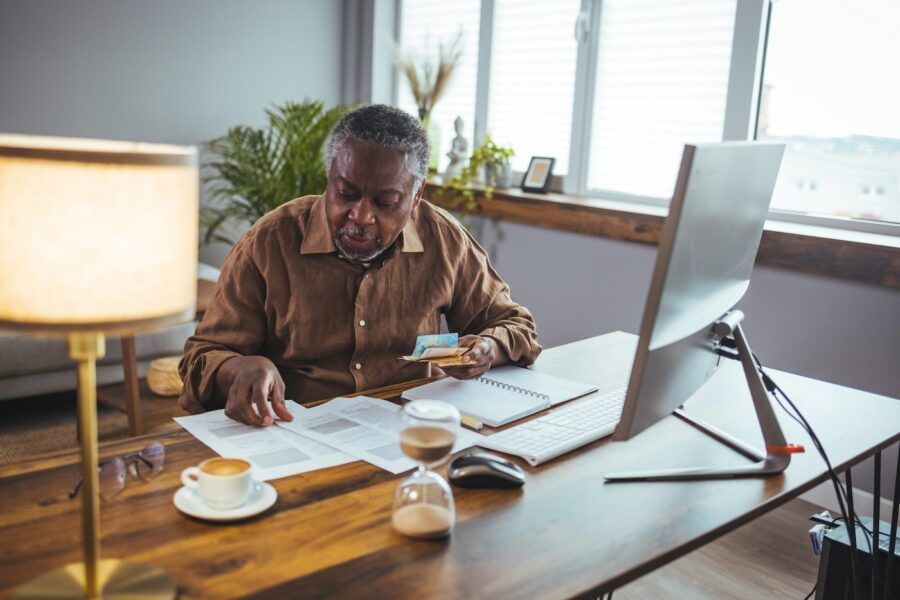 A senior adult man sitting at a table or desk stacked with papers and envelopes working on his home finances.