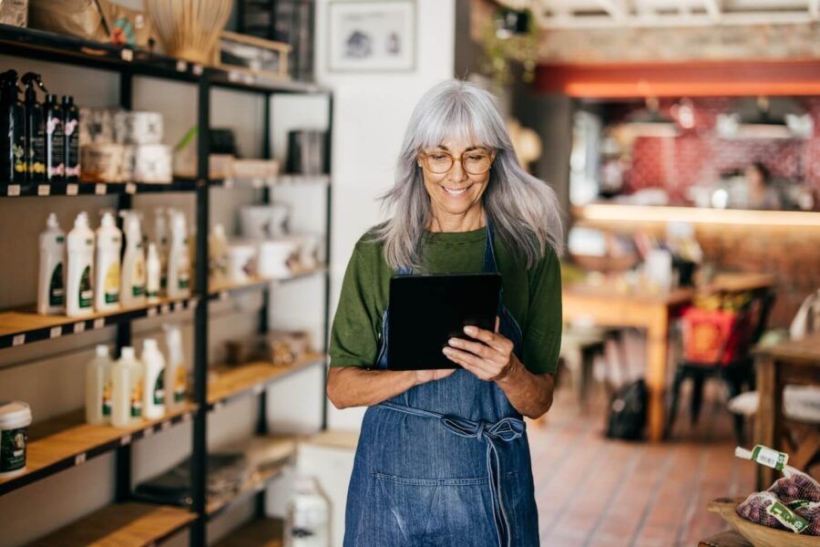 Smiling mature woman wearing an apron stands in a store holding a tablet, with bottles and packaged goods displayed on shelves behind them and a café or shop counter visible further back.