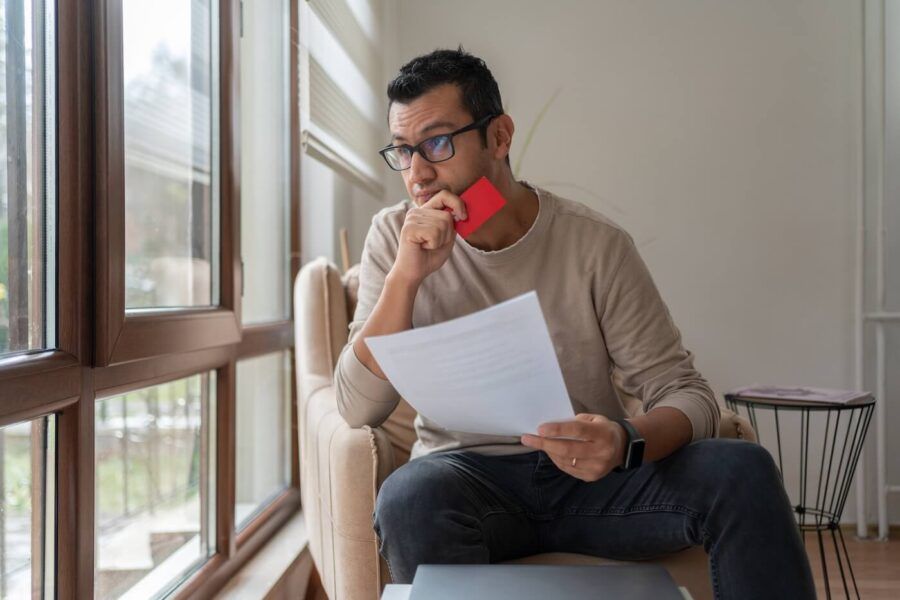 Concerned man sitting in an armchair, looking out the window while holding a credit card and a printout.