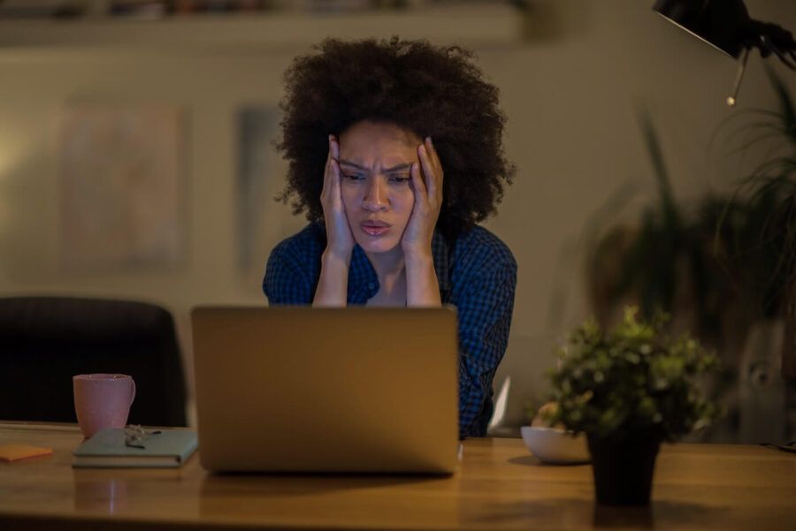 Concerned woman looking at the laptop monitor in a dimly lit room, with both hands placed on the sides of her face. A mug, a notebook, and a small potted plant sit on the desk, while a desk lamp casts warm light and a softly blurred interior is visible in the background.