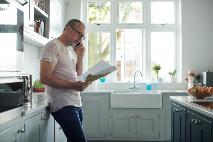 Focused mature man standing in a bright kitchen, reading a printed document while talking on a smartphone