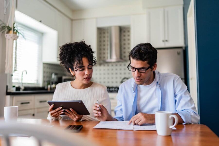 Focused mature couple seated at a kitchen table reviews financial documents together, with one person pointing at paperwork while holding a pen and the other holding a tablet; a mug, a smartphone, and household kitchen elements are visible in the background.