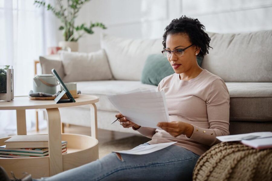 Focused mature woman sitting on the floor next to a coffee table in the living room, reviewing printed documents while filing taxes, with a tablet propped up on the table beside her