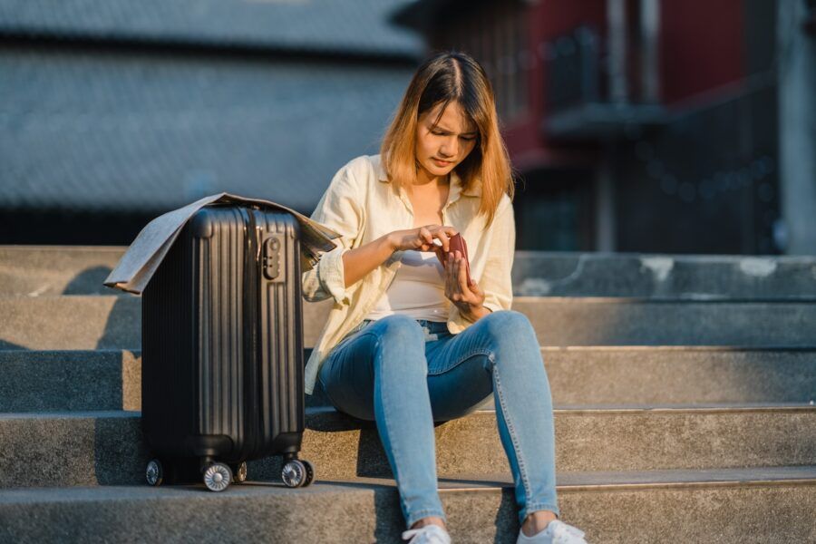 Young woman solo traveler sitting on stairs with suitcase checking her wallet.