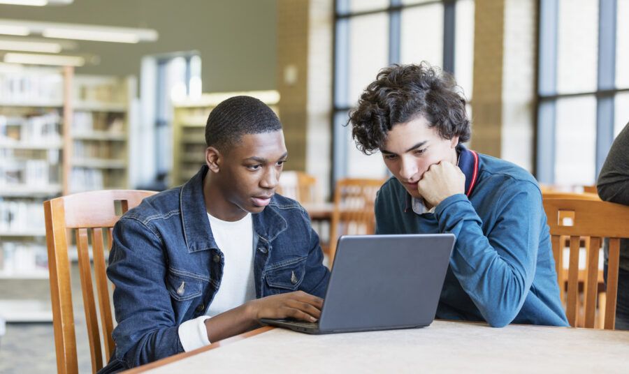 Two teenage boys sitting at a table in the library. One boy is typing on his laptop while his friend watches.