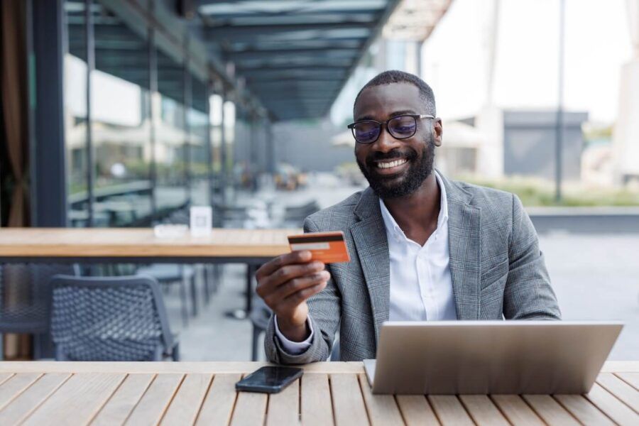 Smiling man in smart casual outfit using a credit card and laptop while seated in an outdoor lounge area
