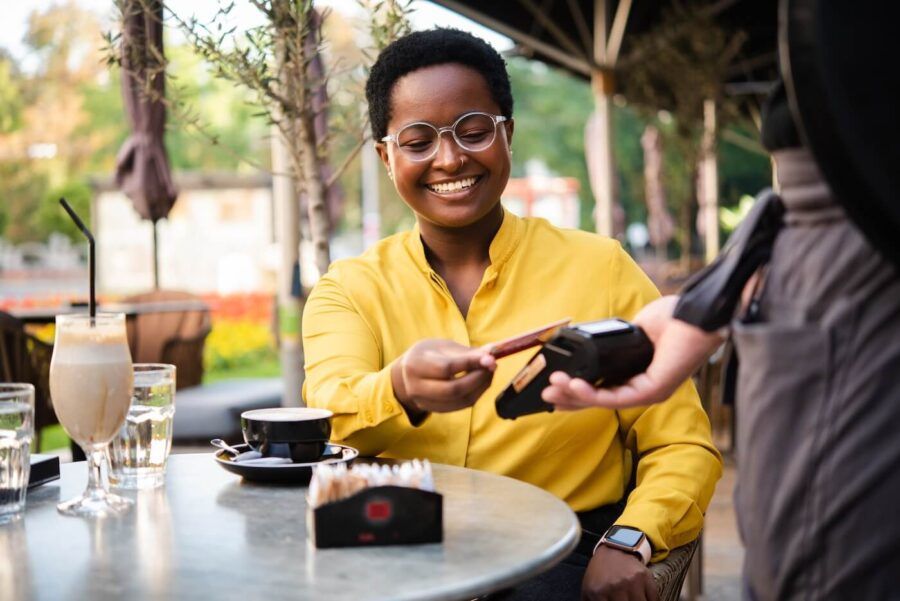 Smiling young woman seated at an outdoor café table hands a payment card to a waiter holding a portable card reader, with drinks, glassware, and a small container on the table, and trees, umbrellas, and café seating visible in the background.