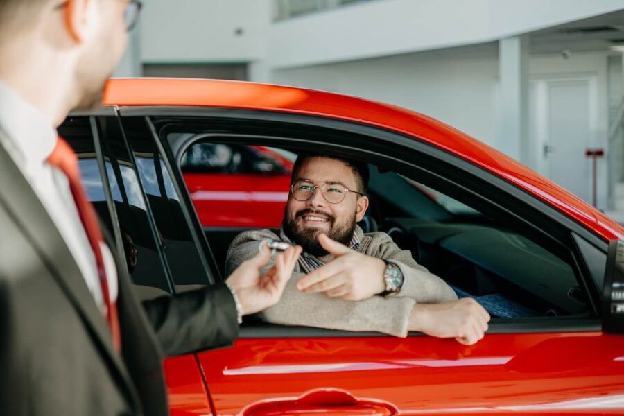 Smiling man sitting in a new orange car, receiving keys from a male dealership agent