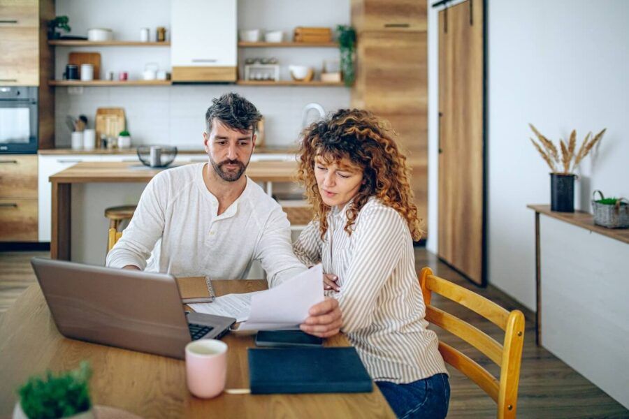 Focused couple sitting at a wooden table in a kitchen, reviewing printed documents together with a laptop open on the table, shelves and kitchen cabinets visible in the background