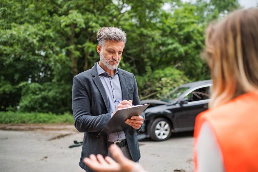 Male insurance adjuster is making a report at at the site of a recent car accident while talking to a female driver