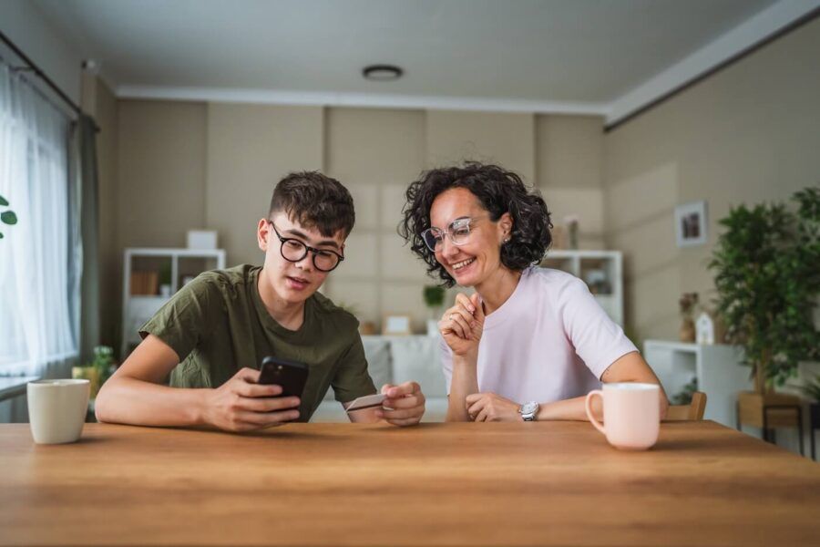 Mature woman and a teenage boy holding a credit card and looking at a smartphone together with mugs on the table