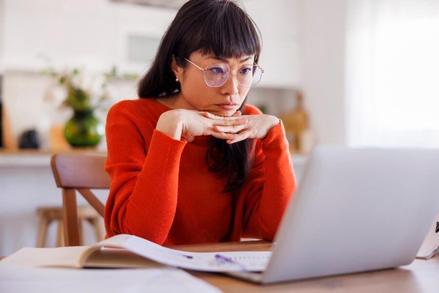 Focused mature woman sits at a table using a laptop, with an open book and papers spread out in front of her in a bright indoor setting.
