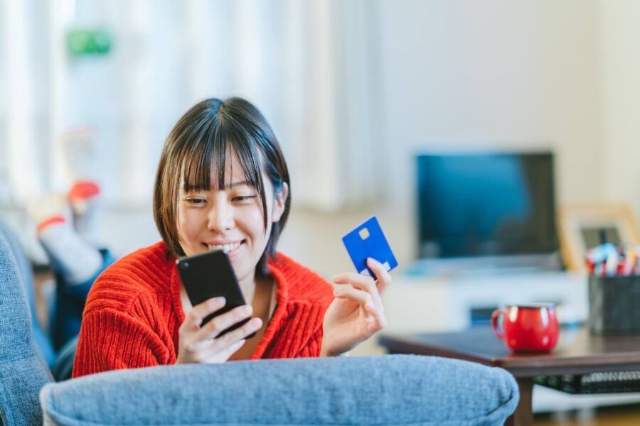 Smiling young woman lying on a sofa with a smartphone and using her credit card