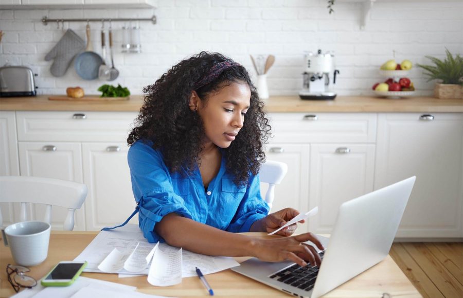 A woman sitting at a table with papers, using her laptop t o work on finances at home.