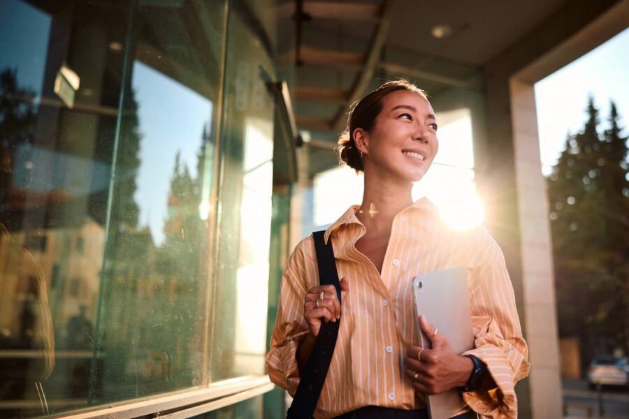 Smiling young woman walking outside a modern office building while carrying a tablet and shoulder bag, with sunlight reflecting off glass windows and trees visible in the background.