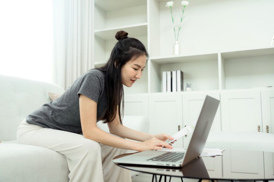 Young woman sits in the living room using her laptop to check her bank account at home.