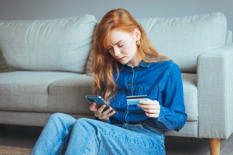 A woman checking her credit card utilization on her phone holding her credit card. She is sitting on the floor of her living room, leaning against the front of her couch.