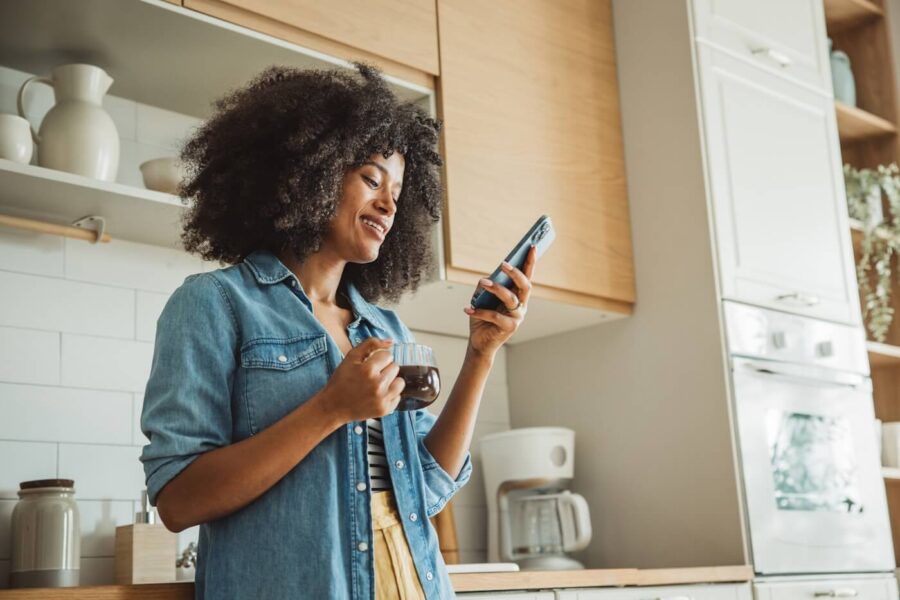 Smiling woman standing in a bright kitchen holding a mug of coffee and looking at a smartphone, with wooden cabinets, open shelving, and a coffee maker visible in the background.