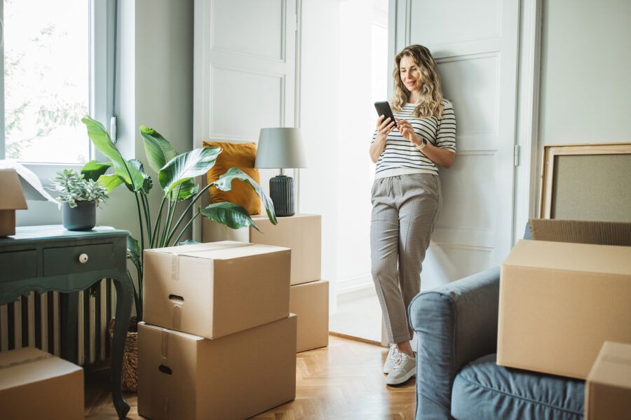 Woman checks her finances on her smartphone in her new home surrounded by moving boxes.