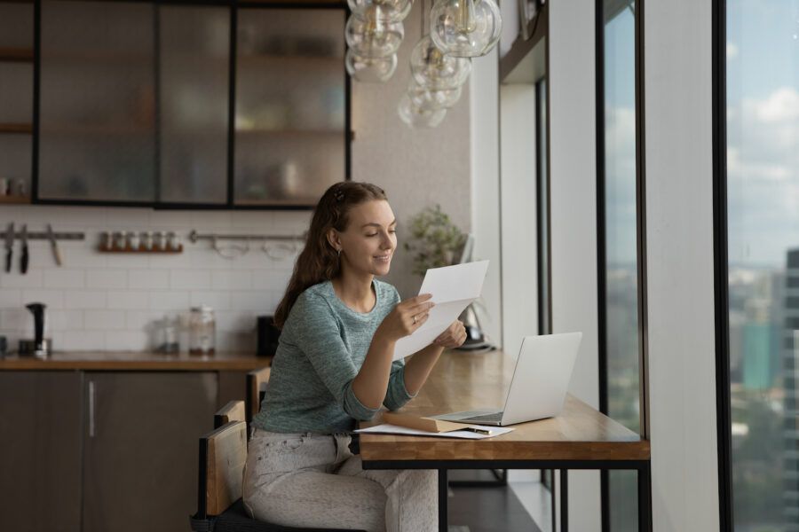 Happy young woman reading positive bank statement at home workplace with laptop.