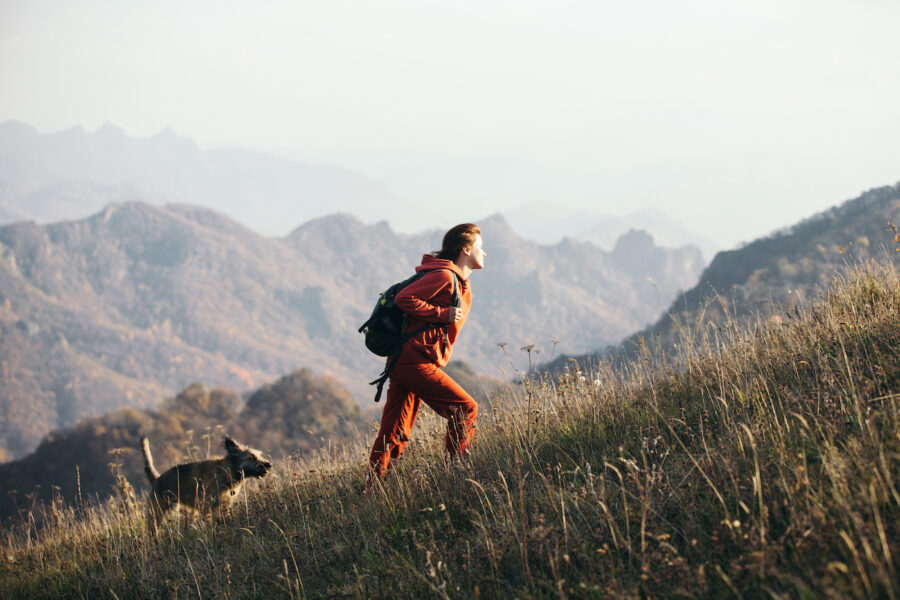 A woman climbs up a grassy mountainside alongside her dog with mountain peaks in the background. She is wearing a backpack and red clothes.