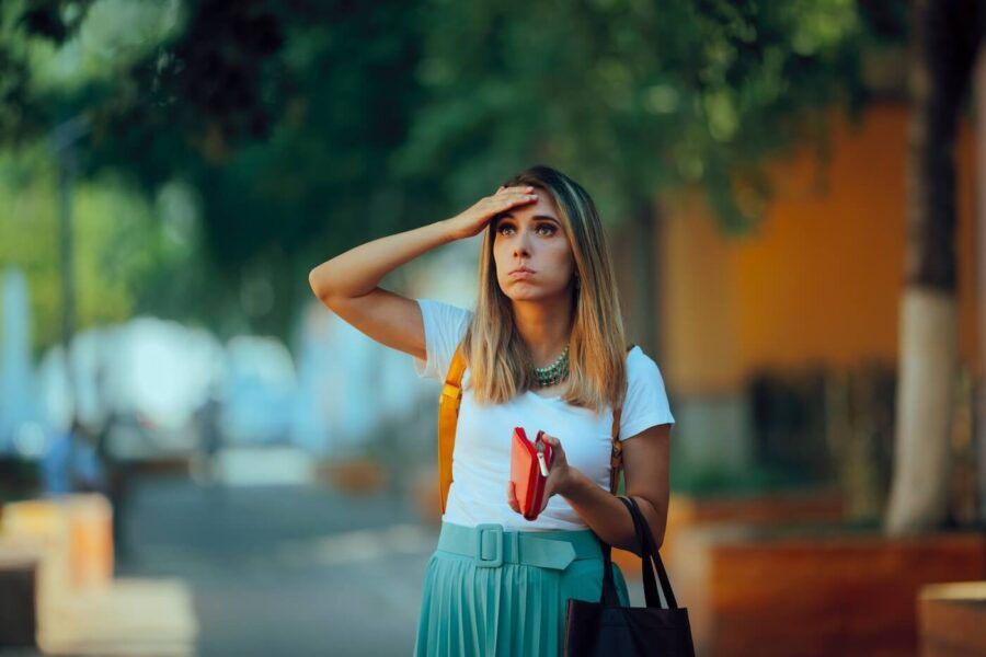 Concerned young woman standing on a tree-lined city street holding an open wallet and a tote bag, with a backpack over one shoulder and residential buildings and greenery in the background.