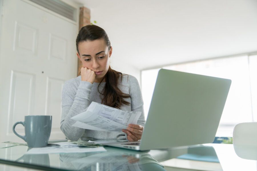 Woman looking worried while considering using her emergency savings fund, looking at paper bills and laptop