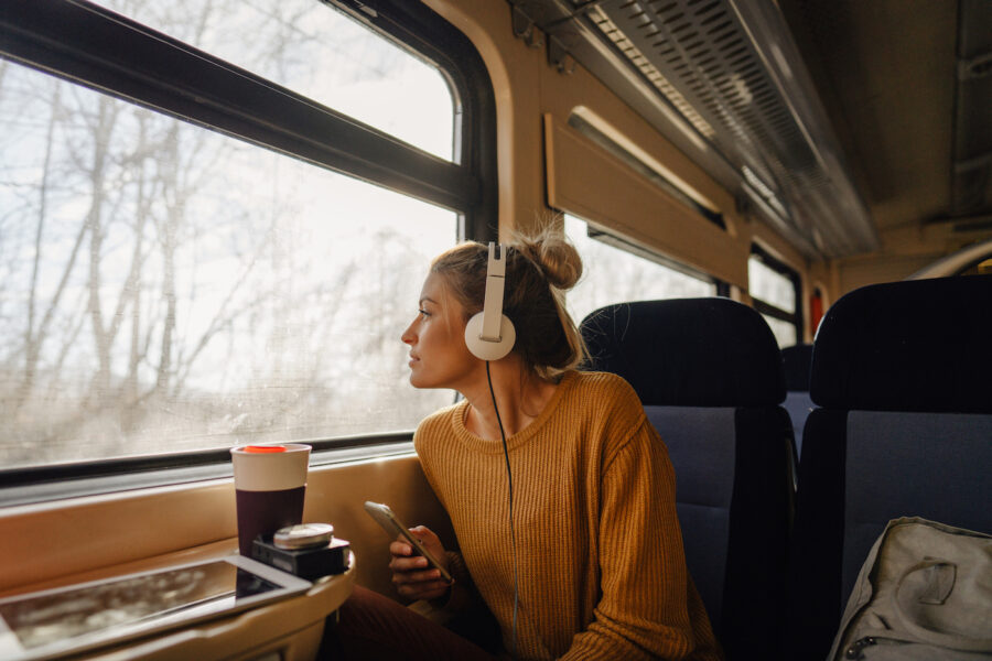 Photo of a young woman riding on a train, enjoying her trip while looking through the window and listening to some music on her mobile phone