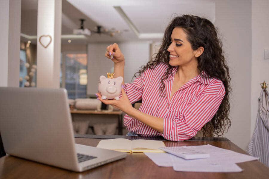 Happy young woman putting money in her piggy bank while sitting at her laptop with papers on her desk, savings concept