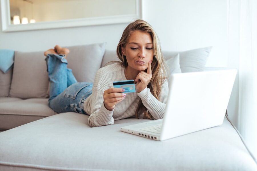 Concerned woman lying on a sofa at home while holding a credit card and using a laptop