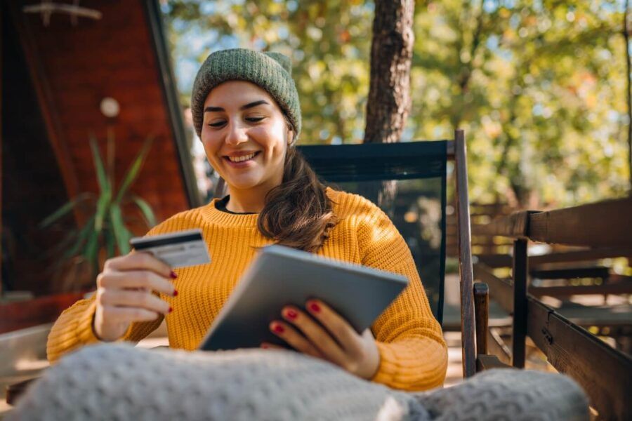 Smiling woman sitting on an outdoor deck holding a credit card and using a tablet, with a cabin-style building and trees in the background, illustrating online shopping or digital banking in a relaxed setting.