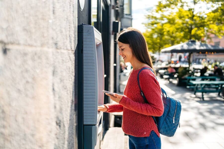 Smiling woman standing at an outdoor ATM, using the machine while holding a smartphone, with a backpack and a city streetscape visible in the background.