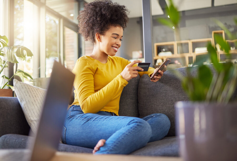 Woman using a credit card and phone sitting on the couch at home