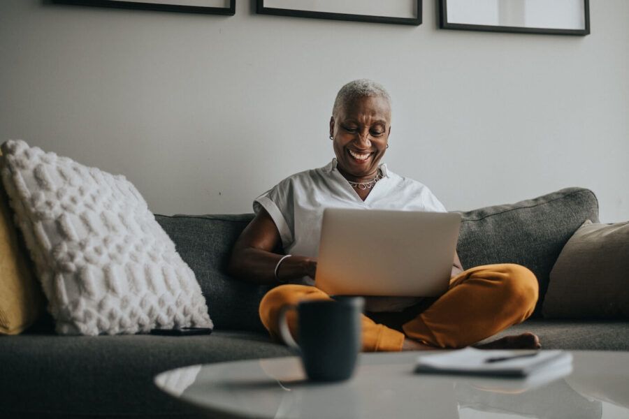 Smiling senior woman sitting cross-legged on a sofa using a laptop in a living room, with a coffee mug on a nearby table, a textured throw pillow, and framed wall art visible in the background.