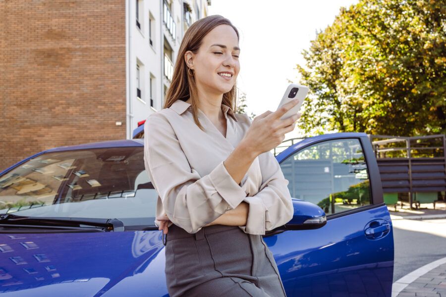 Woman leaning against her car and using a smart phone to get an auto insurance quote.