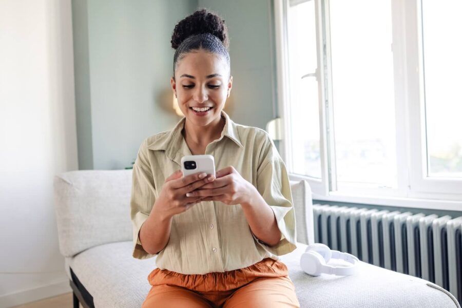 Smiling woman seated on a sofa in a bright living room using a smartphone, with wireless headphones nearby and natural light coming through large windows.